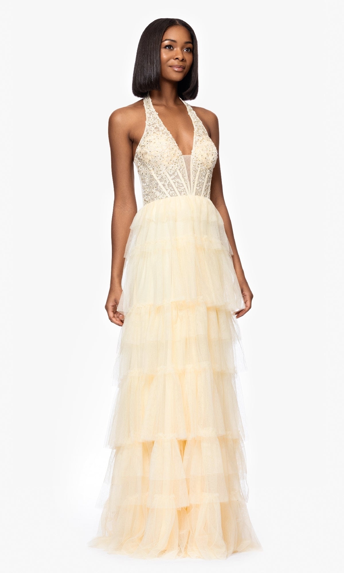A woman wears the Blondie Nites 5349BN, a long yellow halter prom gown featuring a beaded bodice and tiered tulle skirt, radiating charm and elegance.